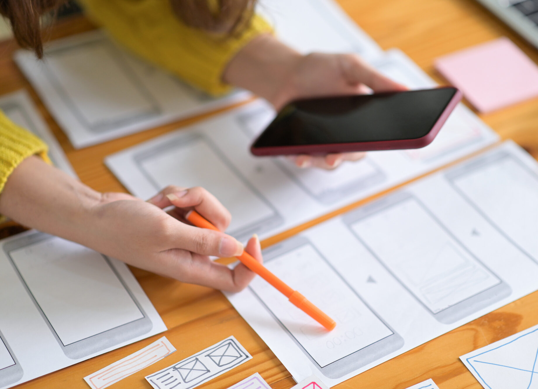 Manos de una persona sosteniendo un smartphone sobre un escritorio lleno de bocetos de wireframe, indicando el desarrollo de apps.